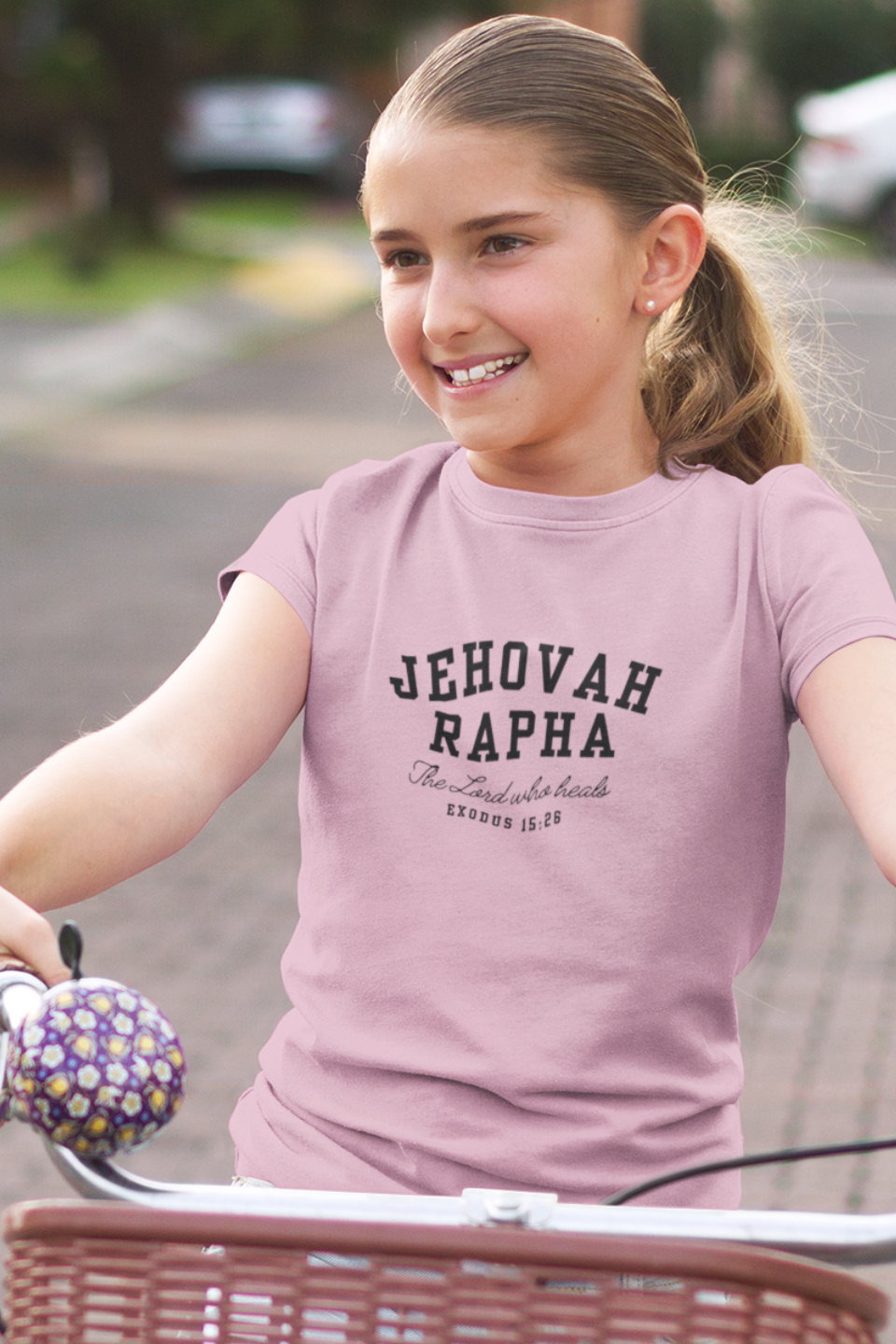 Young girl riding a bike wearing a t-shirt with 'Jehovah Rapha' text.