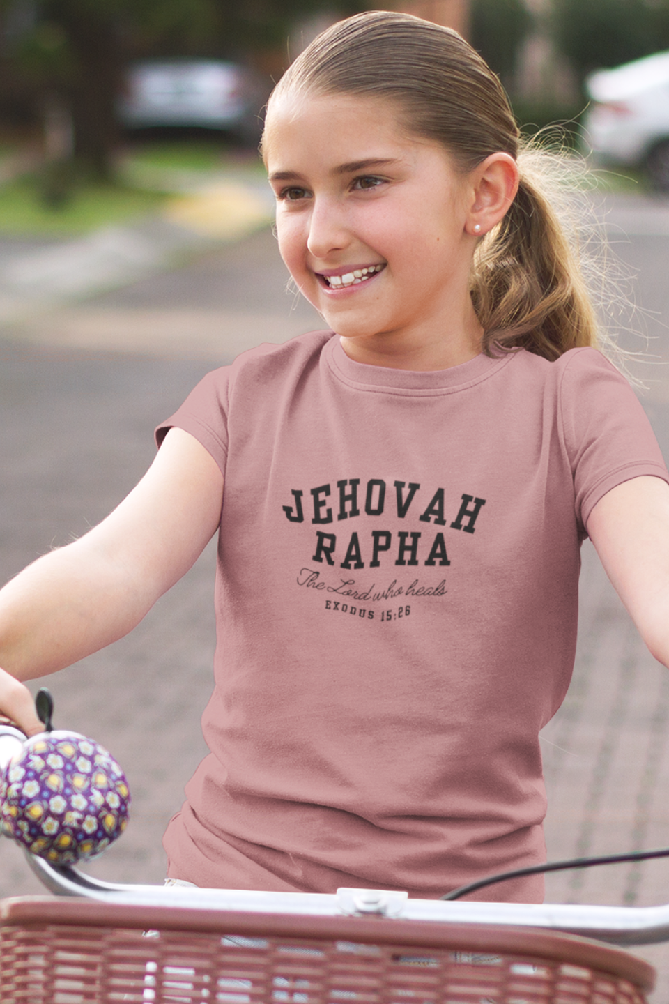 Young girl riding a bike wearing a t-shirt with 'Jehovah Rapha' text.