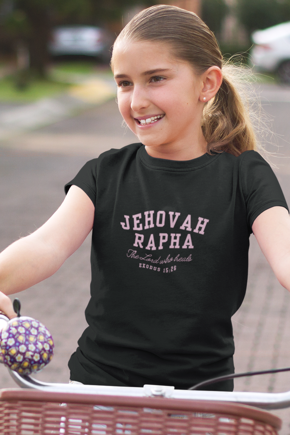 Young girl riding a bicycle with a black 'Jehovah Rapha' t-shirt on a residential street.