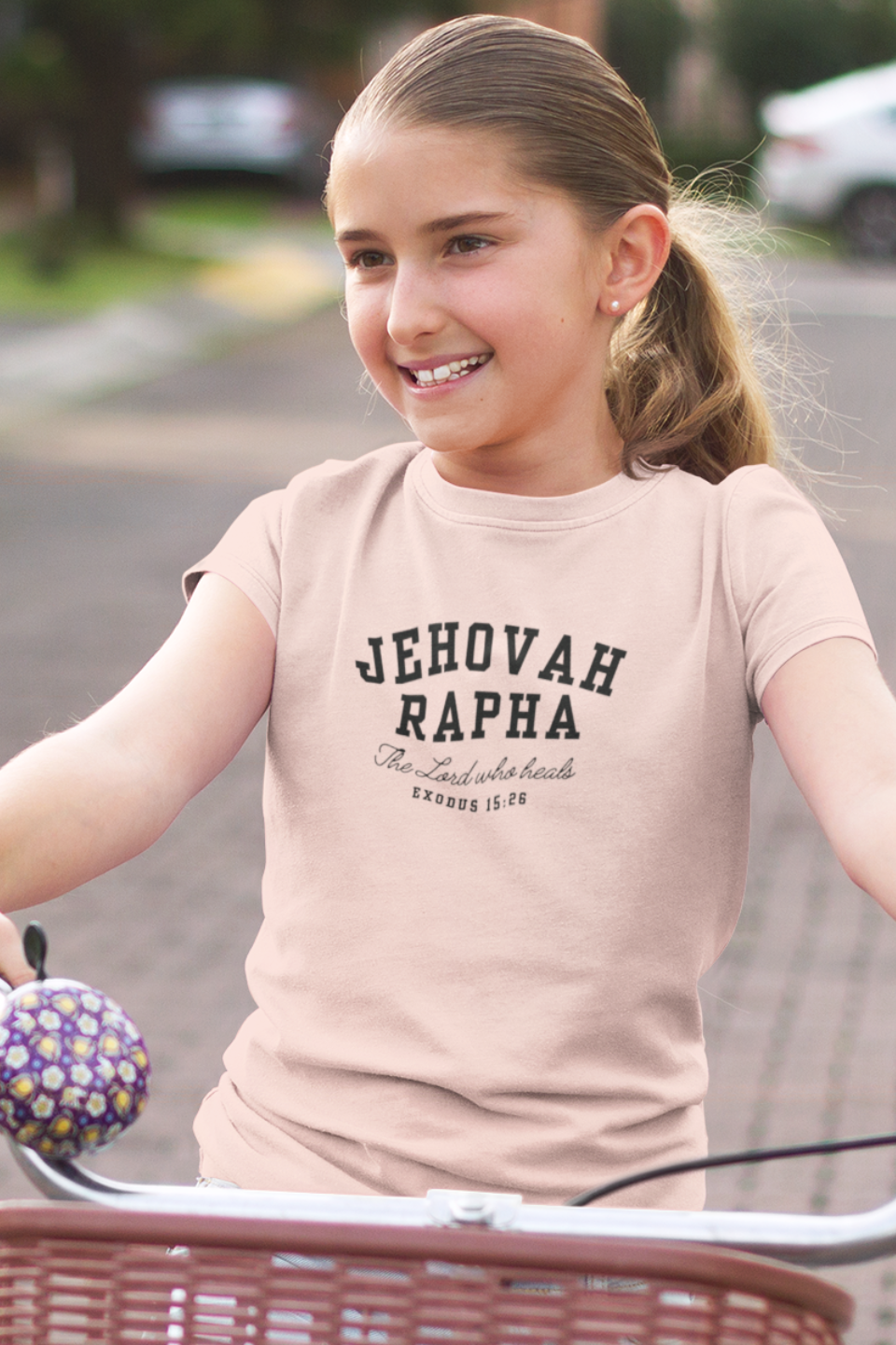 Young girl wearing a peach t-shirt with 'Jehovah Rapha' text, sitting on a bicycle.